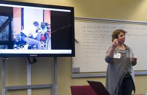 Föreläsaren i grå lång kofta framför en videoskärm.