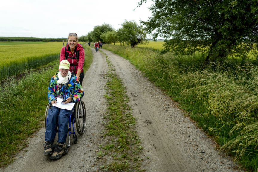 Christina i röd tröja bakom Tony som sitter i rullstol på en grusväg längs vandringsleden.