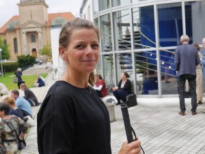 Machteld i ljust hår och hästsvans och märk tröja står utanför en byggnad med denvita käppen i handen,