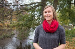 Elin i ljust långt hår, grå klänning och röd sjal står i en park intill en flod.