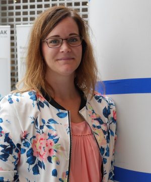 Caroline i halvlångt blont hår och glasögon står, HOn bär en vit jacka med blommor över en rosa blus.