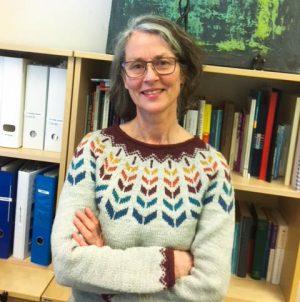 Maj står i färggrann tröja med händerna i kors framför en bokhyylla,. Hon har halvlångt mörkt hår med lite gråa stråk i och glasögon och ler på bilden,