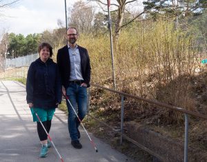 Amanda i mörk jacka, turkos klänning och gympaskor tillvänger och Klas Nelfelt med jeans och kavaj till höger går nerför en backe. båda har rödvit-randig köpp.