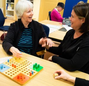 Britt-Marie i kort ljust hår,svart och ljusblårandig tröja ler stort. I höger hand håller hon sin pjäs. Till vänster om henne taktiltolken, I förgrunden Fiaspelet.