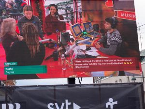 Till vänster står Linda i buren. Intill henne, med ryggen mot kameran står tolken Sra, till vänster token Julia, Farah med flätor tittar på och William som sitter bakom skrivbordet. framför ett antal skärmar och reglage och med hörlurar på sig.