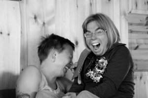 Den svartvita bilden visar två kvinnor som skrattar. Den till vänster har kort mörkt hår, den till höger glasögon och ljust halvlngt hår.