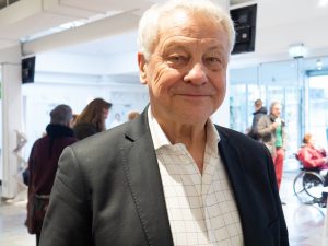 Bengt i mörk kavaj och ljus rutig skjorta,vitt hår och ett leende. I bakgrunden syns andra människor i en fojaé.