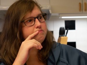 Porträtt. Evelinai brunt halvlångt hår och glasögon tittar åt vänster med fingret mot läppen.