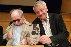 Vilhelm i ljus jacka och rutig skjorta sitter intill Bengt i svart kavja och rutig skjorta. Bengt håller boken i handen