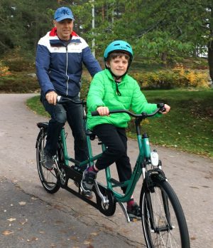 Oliver i grön jacka och cykelhjälm längst fram på tandemcykeln. Pappa är bakom.