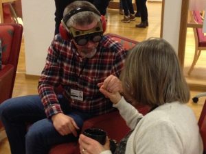 En man i rutig skjorta och jeans med fingerad optik och hörselkåpor håller en kvinna med axellångt hår och ljus ströja i handen. Hon har en kaffekopp i sin andra hand.