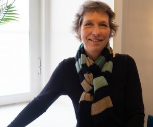 Berit står i ett fönster. Kotklippt hår och svart klänning med en färggrann halsduk virad runt halsen. Hon ler in i kameran.