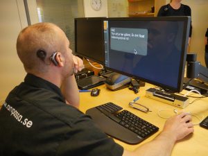 En person med dövblindhet fotad bakfirån. Han har CI och är klädd i svart tröja och sitter framför en dator med förstorad text.