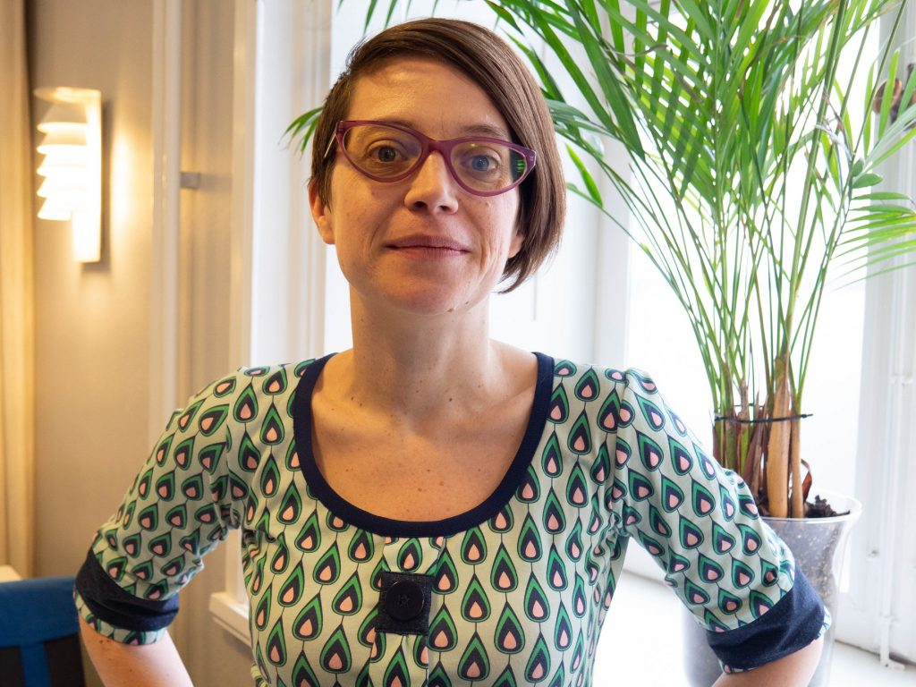 Tove ser bestämd ut och har händerna i midjan. Kort brunt hår, stora glasögomn och en klänning med grafiskt formade dropper i grönt, rosa svart mot en ljusgrön botten.