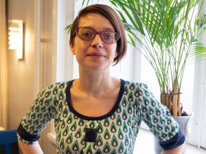 Tove ser bestämd ut och har händerna i midjan. Kort brunt hår, stora glasögomn och en klänning med grafiskt formade dropper i grönt, rosa svart mot en ljusgrön botten.