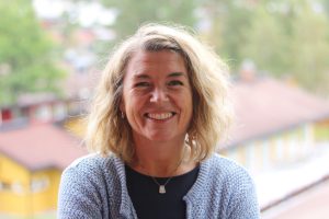 Line med ljust axellångt hår, ljusblå kofta och svart tröja i halvfirur mot en suddig grön bakgrund. Hon ler stort.