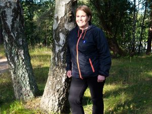 Karen i blå jacka med rött blixtlås står lutad mot en björk i en solig skog.
