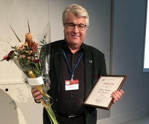 Clase i svart kostym med bomsterbukett i högerhand och diplom i vänster och rödvit namnskylt i blått band runt halsen.
