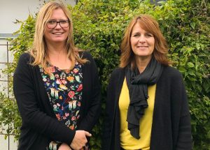 Sofi Malmgren i ljust halvlångt hår och glasögon, blommig klänning och svart trja. Jessica till höger med brunt hår till axlarna, gul tröja, svart schal och svart tröja. De står framfr en grön buske.