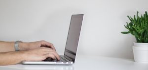 två händer som arbetar vid en laptop som står mittemot en grön växt i kruka