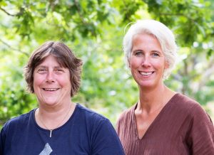 Karin till vänstr i kort, brunt hår och blå tröja. jenny till höger, ljust hår, brunt V-ringat plagg mot en bakgrund av gröna träd.