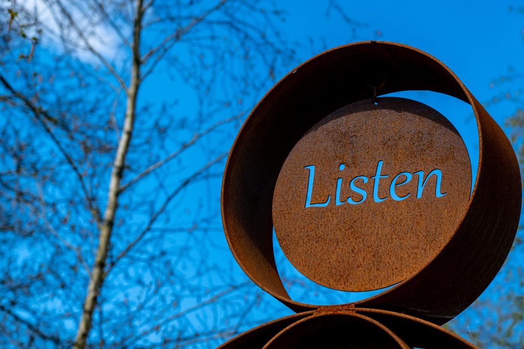 Solbelyst metallskulptur med ordet "Listen" utskuret med en björk och blå himmel i fonden., foto