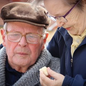 Närbild på en äldre man och kvinna. Båda bär glasögon, mannen har en brun keps och kvinnan håller i något fyrknatigt vitt som båda tittar på. Foto.