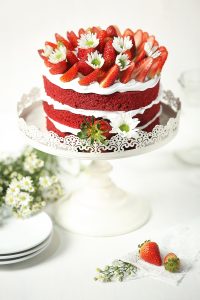 Hög jordgubbstårta på ett vitt tårtfat på fot som står på en vit duk intill några staplade assietter och en liten bukett med prästkragar. Foto från Pixabay.