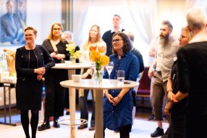 Lena Göransson, iklädd marinblå klänning, står vid ett ståbord omgiven av massa människor. Foto.