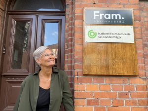 Helene Engh tittar leende upp mot himmeln. På tegelväggen bredvid henne hänger två skyltar. En där det står "Fram Advokatbyrå" och en där det står "Nationellt kunskapscenter för dövblindfrågor". Foto.