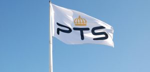 En vit flagga med PTS logga fladdrar i vinden framför en blå himmel. Foto.