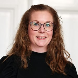 Porträttbild på Emy Hurtig. Hon ler in i kameran och bär svarta glasögon och svart tröja. Foto.