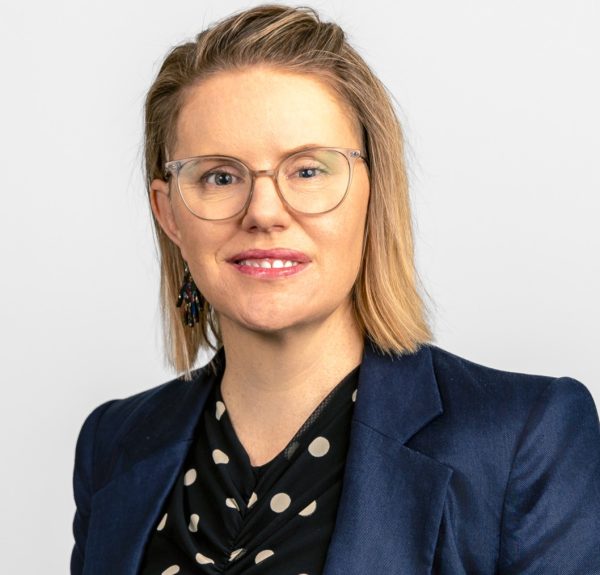 Porträttbild på Hannah McPierzie som bär en svart- och vitprickig topp, en mörkblå kavaj, färgglada örhängen och glasögon med ljus båge. Foto.