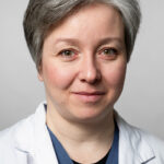 En porträttbild på Katarzyna Mascianica. Hon har grått, kort hår, bär läkarrock och tittar leendes in i kameran. Foto.
