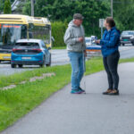 Två personer står och pratar på en trottoar. Den ena personen har en vit käpp i handen. På vägen bredvid dem kör trafik i båda riktningarna. Foto.