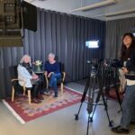 Två kvinnor sitter på var sin stol framför två kameror. Tre lampor är riktade mot dem. Bakom kamerorna står en kvinna och håller i en av kamerorna. De tre kvinnorna tittar alla in i kameran som tar bilden och ler. Foto.