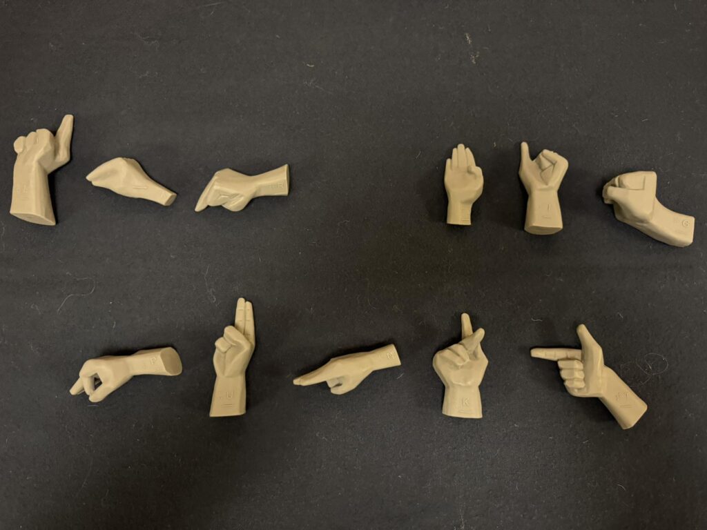 Plasthänder där varje hand gör en av bokstäverna i handalfabetet ligger på en svart yta. Händerna stavar ut "Lär dig punkt". Foto.