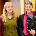 Silje Larsen och Lynn Skrei står bredvid varandra. Silje har blont hår, grön tröja och röd kofta, Lynn har svart kostym och rosa scarf. Båda har glasögon satta på huvudet. Båda ler in i kameran. Lynn bär en bukett blommor. Foto.