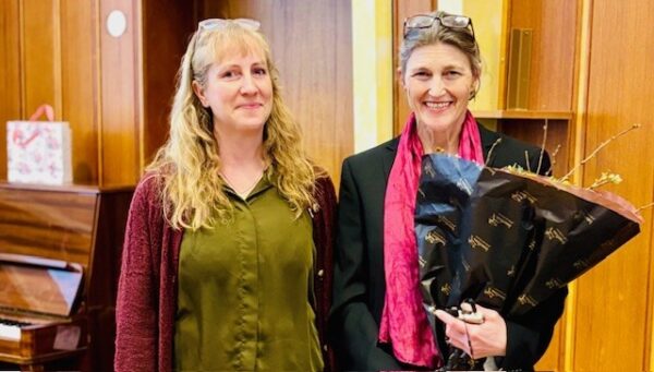 Silje Larsen och Lynn Skrei står bredvid varandra. Silje har blont hår, grön tröja och röd kofta, Lynn har svart kostym och rosa scarf. Båda har glasögon satta på huvudet. Båda ler in i kameran. Lynn bär en bukett blommor. Foto.