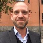 Porträttbild på Filip Asp. Han bär kavaj och skjorta, har skägg och rakat huvud. Han ler in i kameran. Foto.