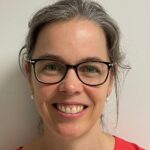 Porträttbild på Julie Defour. Hon bär glasögon, pärlörhängen, en röd tröja och har uppsatt hår. Hon ler in i kameran. Foto.