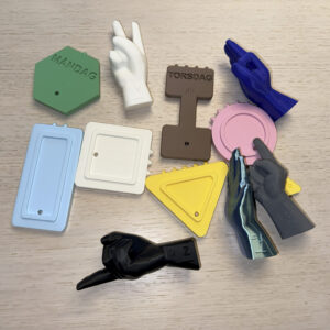 Ett foto av olika 3D-utskrivna material. Här syns fram och baksidor på olika veckodagssymboler samt händer som visar handtecken för v, h, z, j och l.