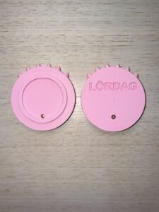 Symbolen för lördag. Formen är en cirkel med 6 taggar på toppen. På baksidan står i relief Lördag och i punkt l. Färgen är rosa. Foto