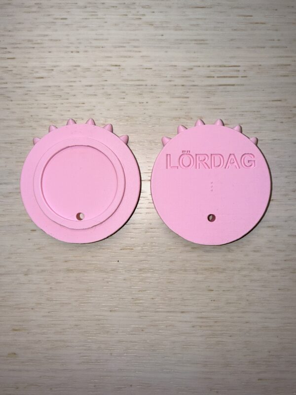 Symbolen för lördag. Formen är en cirkel med 6 taggar på toppen. På baksidan står i relief Lördag och i punkt l. Färgen är rosa. Foto