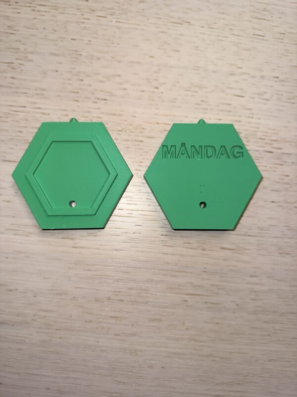 Symbolen för måndag. Formen är liksidigt sexkantig med en tagg på toppen. På baksidan står i relief Måndag och i punkt m. Färgen är grön. Foto