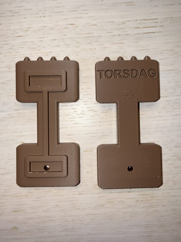 Symbolen för torsdag. Formen är bred uppe och nere och smalare på mitten med 4 taggar på toppen. På baksidan står i relief Torsdag och i punkt to. Färgen är brun. Foto